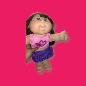 May include: A doll with dark brown hair and blue eyes, wearing a light pink short-sleeved shirt featuring a purple heart graphic with the text CPK, paired with a purple ruffled skirt. The doll has a light skin tone and a smiling facial expression.