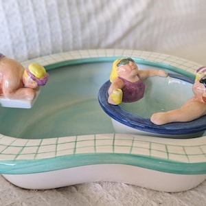 Può includere: Un piatto in ceramica a forma di piscina con tre statuette. Una statuetta sta per tuffarsi, un'altra è in acqua e la terza si sta rilassando. Il piatto ha una base bianca, un interno turchese e un bordo a piastrelle verdi e bianche.