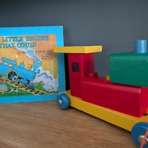 Könnte beinhalten: Ein farbenfroher Holzspielzeugzug mit roten, gelben, grünen und blauen Elementen steht neben einem Kinderbuch mit dem Titel "The Little Engine That Could" von Watty Piper. Das Buch hat einen blauen Einband mit einer Zugillustration.