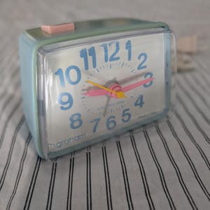May include: A vintage light blue alarm clock with a clear plastic cover. The clock face has blue numerals, pink hands, and a yellow second hand. The clock is approximately 4 inches wide and has a pink button on top.