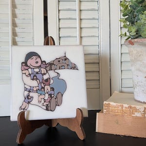 May include: A square white ceramic tile featuring a colorful illustration of a large figure carrying multiple smaller figures on their back and arms, with a stylized mountain landscape in the background, displayed on a small wooden easel stand.