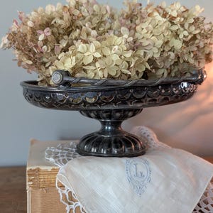 Può includere: Una coppa su piedistallo in metallo argentato con bordo smerlato e manico intrecciato, riempita di fiori di ortensia essiccati. Sotto la coppa si trova un fazzoletto di stoffa bianca con una lettera L ricamata in blu e un centrino all'uncinetto.