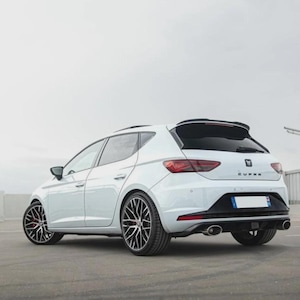 Può includere: Un'auto hatchback Cupra bianca vista da un'angolazione di tre quarti posteriore. Il veicolo presenta cerchi in lega scuri a razze multiple, pinze dei freni rosse, un diffusore posteriore nero con doppio scarico e il logo Cupra sul portellone.