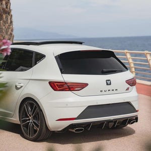 以下が含まれることがあります： 海辺に駐車された、黒いリムとサンルーフを備えた白いハッチバック車。車の後部には「CUPRA」の文字と赤い「S」エンブレムがあります。車のテールライトは赤色です。