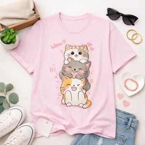 May include: A light pink t-shirt featuring a stack of three cartoon cats. The top cat wears a floral crown, the middle cat holds a heart, and the bottom cat smiles. The words "Wow!!" and "Hi" are printed on the shirt.