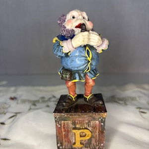 May include: A hand-painted figurine of a character with purple hair, wearing a blue jacket with yellow trim and brown pants, standing on a wooden block base. The base features a prominent gold-colored letter P on the front and a wood grain texture.