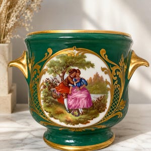 May include: A deep green ceramic vessel featuring two gold-toned handles and a central circular medallion. The medallion depicts a painted scene of two figures in a landscape setting. The rim and base are accented with gold trim and ornate gold filigree.