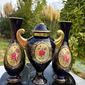 May include: A set of three dark blue ceramic pieces with gold trim and floral motifs. Two tall vases flank a central lidded urn with two gold handles. Each piece features an oval medallion containing a bouquet of red roses and small blue flowers.