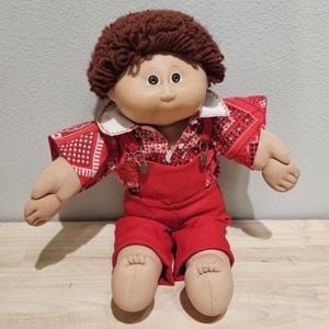 Pode incluir: Uma boneca de pano com cabelo de lã castanho e rosto redondo. A boneca veste uma camisa vermelha com colarinho branco e um padrão de bandana branco e preto, combinada com jardineiras vermelhas lisas com fivelas metálicas nas alças.