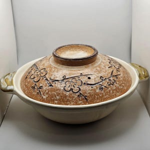 May include: A round ceramic cooking pot with a lid, featuring a speckled tan and cream finish. The lid is decorated with dark brown hand-painted floral branch motifs. The pot has two side handles with green and brown glaze accents.