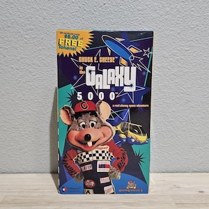 Può includere: Una copertina di videocassetta VHS per Chuck E. Cheese in the Galaxy 5000, a real cheesy space adventure. La copertina presenta un personaggio topo con un berretto rosso e una tuta da corsa, un'astronave blu e grafiche a stella su sfondo viola.