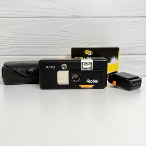 May include: A black Rollei A110 subminiature camera with a silver lens and orange accents. The camera is accompanied by a black leather case, a small black flash attachment with an orange button, and a yellow and black box labeled E3.