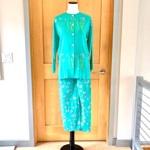 May include: A turquoise two-piece outfit on a mannequin. The long-sleeved top has a button-up front and floral accents. The matching pants feature a white floral pattern. The outfit is displayed against a wooden door.