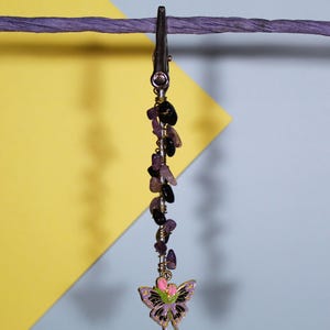 May include: A metal alligator clip featuring a vertical wire wrapped with small, tumbled purple amethyst and black obsidian stone chips. A butterfly charm with purple, black, and pink enamel details hangs from the bottom of the wire structure.