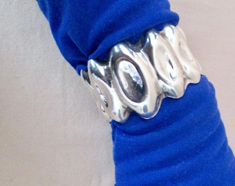 May include: A silver cuff bracelet with an abstract design is worn over a blue sleeve. The bracelet has a series of oval shapes linked together. The sleeve is a vibrant blue color.