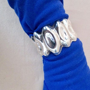 May include: A silver cuff bracelet with an abstract design is worn over a blue sleeve. The bracelet has a series of oval shapes linked together. The sleeve is a vibrant blue color.