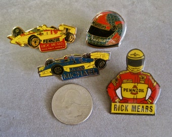 May include: Four enamel pins featuring race car drivers and logos. The pins feature a yellow race car with the words "Pennzoil Rick Mears", a blue race car with the word "Michael", a red and green helmet with the word "Marlboro", and a driver in a red and yellow racing suit with the words "Pennzoil Rick Mears".