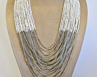 May include: A multi-strand necklace made of small white and silver beads. The necklace is layered and hangs down the front of the neck.