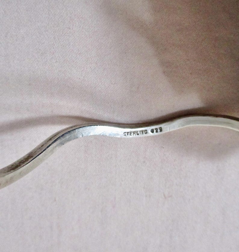 May include: Close-up of a silver, wavy bracelet. The bracelet is stamped with "STERLING 925". The silver has a polished finish. The bracelet is on a neutral background.