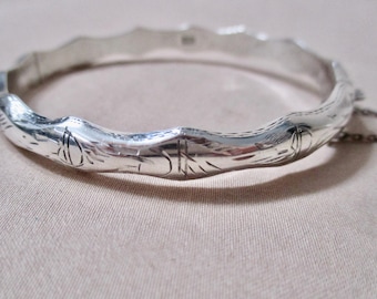 May include: A silver-colored bangle bracelet with a textured, wavy design. The bracelet has etched floral and geometric patterns. The clasp is connected to a small chain. The bracelet is made of a shiny metal.