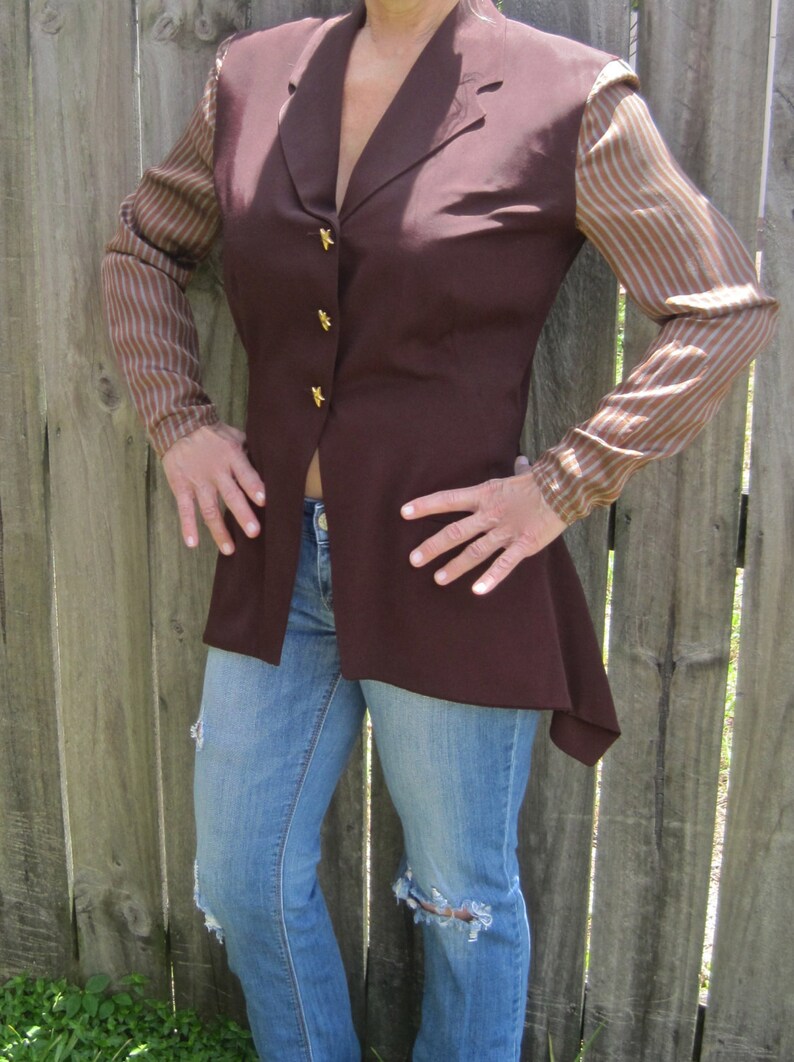 May include: A brown blazer with a long, asymmetrical hemline. The blazer has gold buttons and striped sleeves. The blazer is worn over a pair of blue jeans.