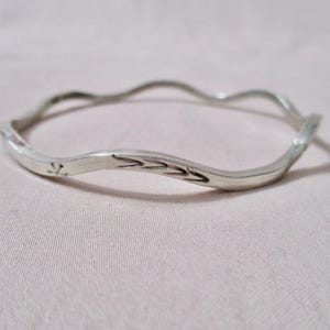 May include: A silver-colored bangle bracelet with a wavy design. The bracelet has a smooth, polished finish and features etched decorative elements. The bracelet is a simple, elegant piece of jewelry.