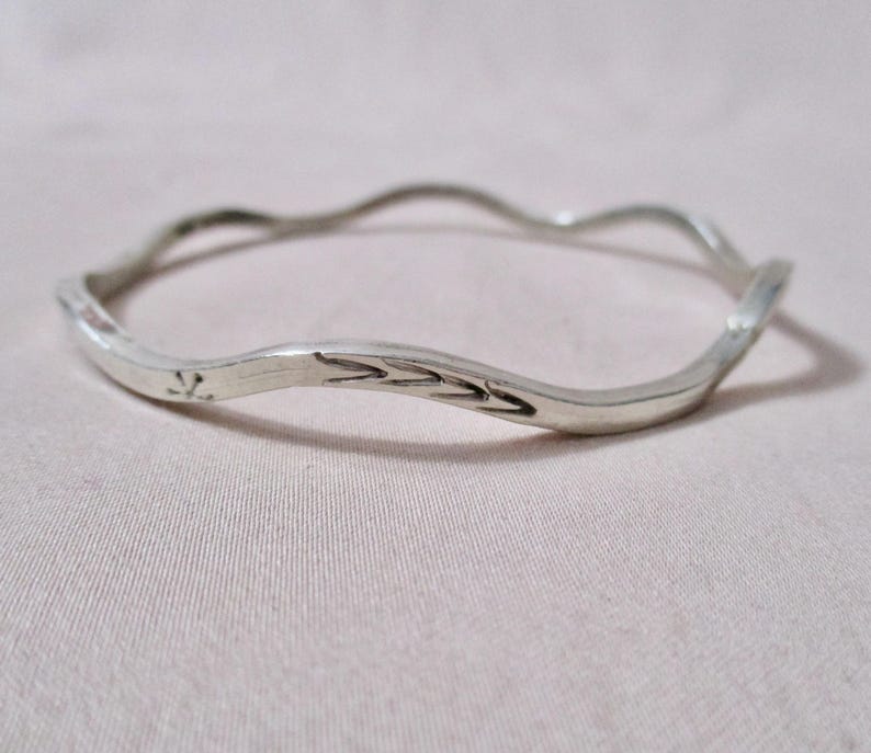 May include: A silver-colored bangle bracelet with a wavy design. The bracelet has a smooth, polished finish and features etched decorative elements. The bracelet is a simple, elegant piece of jewelry.