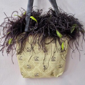 May include: A small, beige handbag with black leather straps and a black and green ostrich feather trim. The bag has a repeating pattern of monkeys on the front.