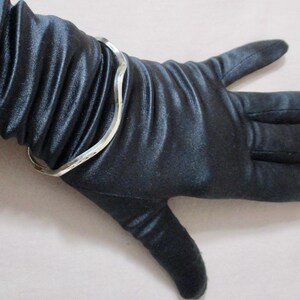 May include: A silver-colored, wavy-design bracelet on a black satin glove. The bracelet has a simple, elegant design. The glove is smooth and shiny, with visible folds in the fabric. The overall aesthetic is one of sophistication and style.