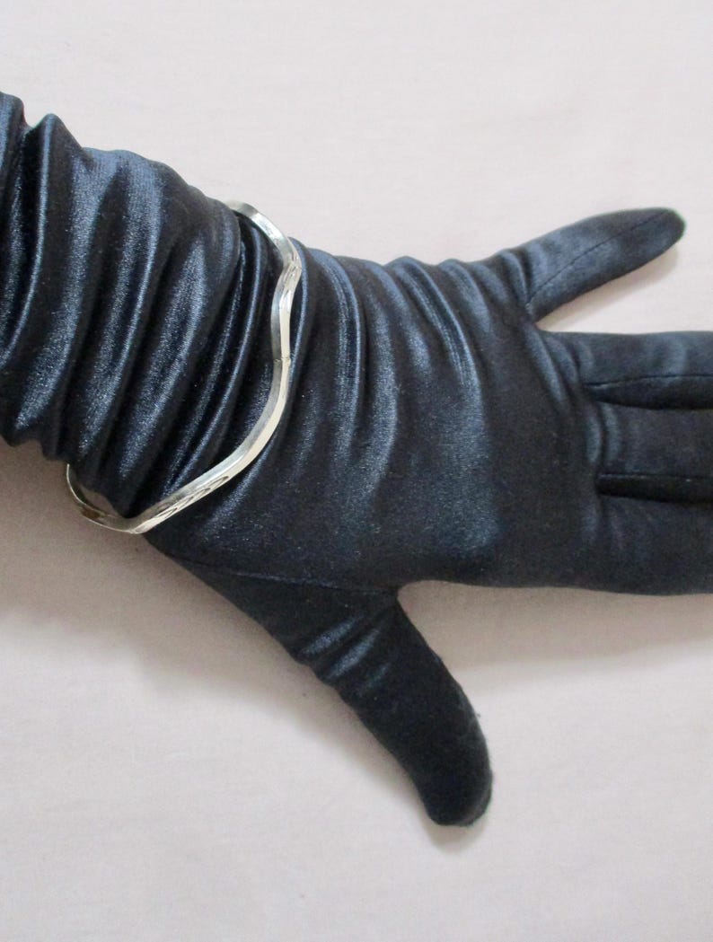 May include: A silver-colored, wavy-design bracelet on a black satin glove. The bracelet has a simple, elegant design. The glove is smooth and shiny, with visible folds in the fabric. The overall aesthetic is one of sophistication and style.