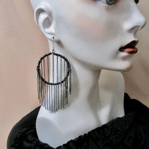 May include: Black hoop earrings with dangling chains. The earrings are attached to a mannequin's ear. The hoop is lined with small black beads. The chains hang down from the hoop, creating a fringe effect. The mannequin is wearing a black top.