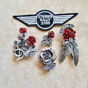 May include: Five motorcycle rally pins with red roses and silver accents. The pins feature the text "Daytona 1999", "Daytona 98", "Daytona 93", "Sturgis Rally 2000", and "Sturgis 97".