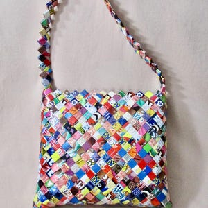 May include: A colorful, woven handbag with a shoulder strap. The bag is made from small, square pieces of paper in various colors and patterns, creating a patchwork design. The strap is also woven from the same materials.