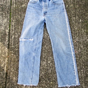 May include: A pair of light blue denim jeans with a white seam down the outside of each leg. The jeans have a distressed look with a small rip on one leg and frayed hems.