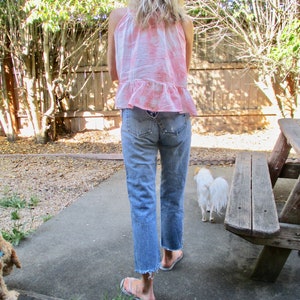 May include: A woman wearing a pink and white tie-dye tank top with a ruffled hem and light blue denim jeans. She is walking with a small white dog in a backyard setting.