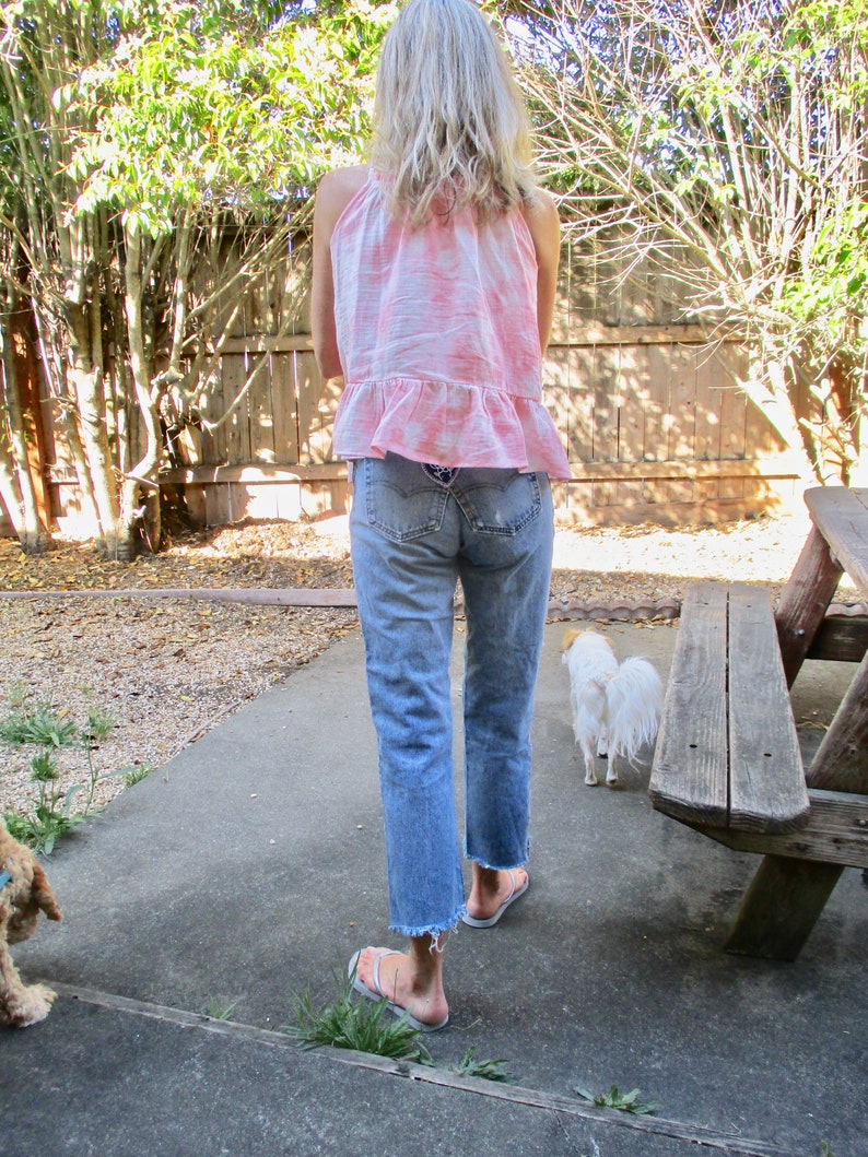 May include: A woman wearing a pink and white tie-dye tank top with a ruffled hem and light blue denim jeans. She is walking with a small white dog in a backyard setting.