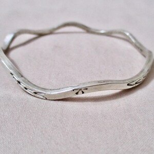 May include: A silver-toned, wavy bangle bracelet. The bracelet has a textured, irregular surface with etched floral designs and a small flower-like emblem. The bracelet is a simple, yet elegant piece of jewelry.