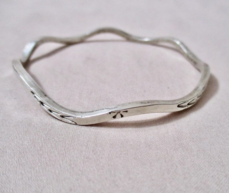 May include: A silver-toned, wavy bangle bracelet. The bracelet has a textured, irregular surface with etched floral designs and a small flower-like emblem. The bracelet is a simple, yet elegant piece of jewelry.