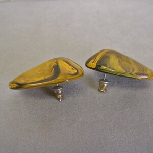 May include: A pair of abstract stud earrings. Each earring has a rounded, triangular shape with a post back. The earrings feature a marbled pattern in shades of yellow and brown, with a glossy finish. The earrings are set against a neutral background.