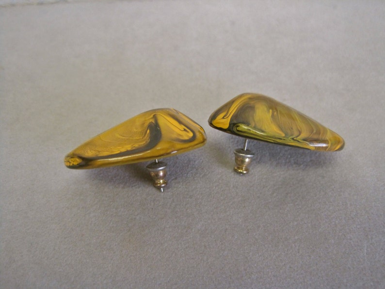 May include: A pair of abstract stud earrings. Each earring has a rounded, triangular shape with a post back. The earrings feature a marbled pattern in shades of yellow and brown, with a glossy finish. The earrings are set against a neutral background.