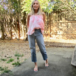 May include: A woman wearing a pink tie-dye tank top and blue jeans with distressed details. She is standing on a concrete patio with a wooden fence and green plants in the background.