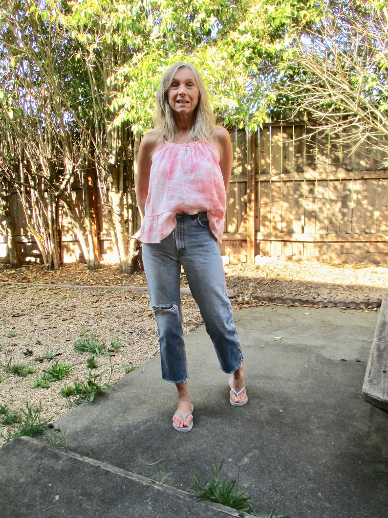 May include: A woman wearing a pink tie-dye tank top and blue jeans with distressed details. She is standing on a concrete patio with a wooden fence and green plants in the background.