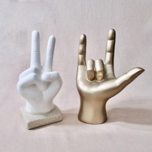 May include: Two hand sculptures: one white, gesturing the peace sign, and one gold, forming the "I love you" sign. The white hand is on a speckled base. The gold hand is smooth and shiny. Both are decorative.