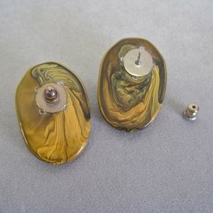May include: A pair of oval-shaped stud earrings with a swirling pattern of gold, olive green, and brown. The earrings have a post and backing for secure wear. The design is reminiscent of marbled stone.