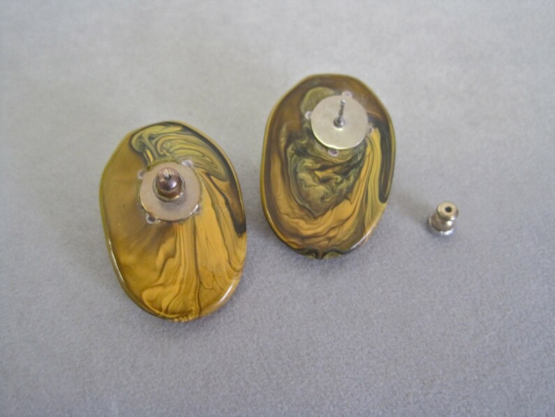May include: A pair of oval-shaped stud earrings with a swirling pattern of gold, olive green, and brown. The earrings have a post and backing for secure wear. The design is reminiscent of marbled stone.