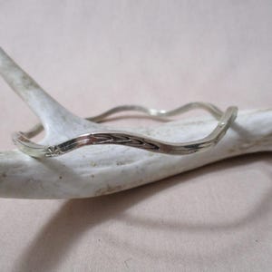 May include: A silver-colored bangle bracelet with a wavy design. The bracelet has a textured surface and is resting on a white antler. The bracelet's design includes a small floral pattern.
