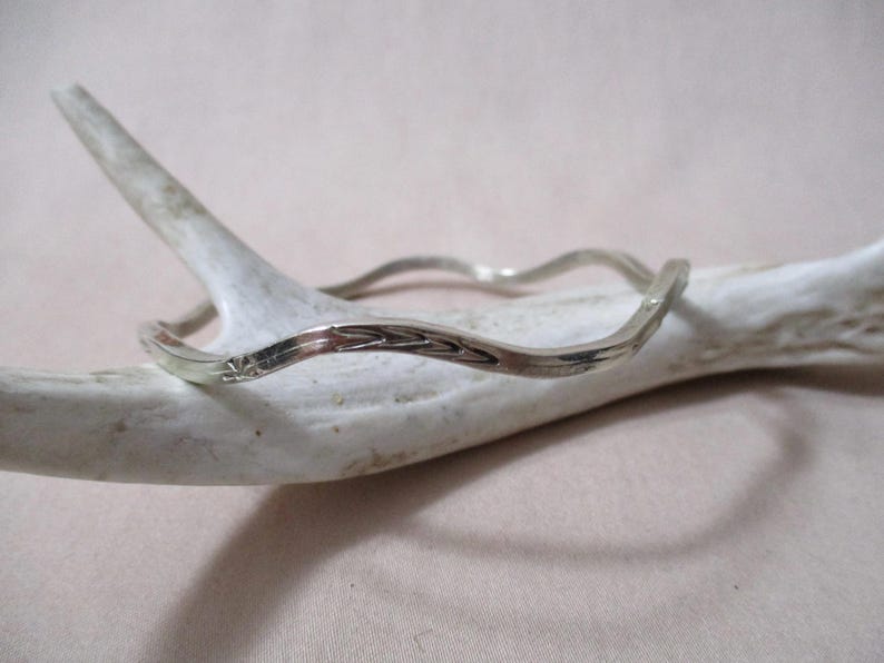 May include: A silver-colored bangle bracelet with a wavy design. The bracelet has a textured surface and is resting on a white antler. The bracelet's design includes a small floral pattern.
