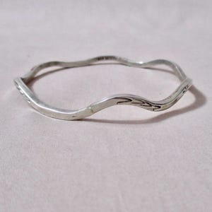 May include: A silver-colored bangle bracelet with a wavy, organic design. The bracelet has a textured surface and appears to be made of metal. The overall style is minimalist and elegant, suitable for everyday wear.