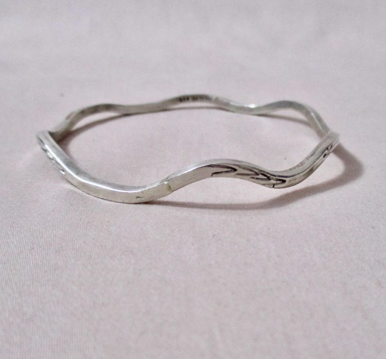 May include: A silver-colored bangle bracelet with a wavy, organic design. The bracelet has a textured surface and appears to be made of metal. The overall style is minimalist and elegant, suitable for everyday wear.