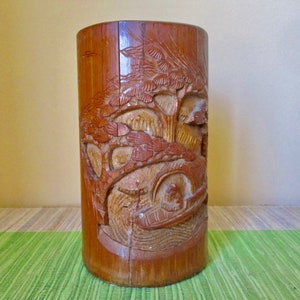 May include: A brown bamboo vase with a carved design of a tree and a boat. The vase is sitting on a green mat.